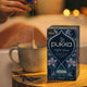 Pukka night time tea box with a steaming cup of tea and lavender on a wooden surface.
