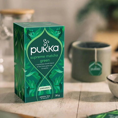 Box of Pukka Herbs Supreme Matcha Green Tea with a blurred background of tea brewing.