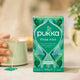 Pukka three mint tea box on a blurred background with a hand holding a cup.