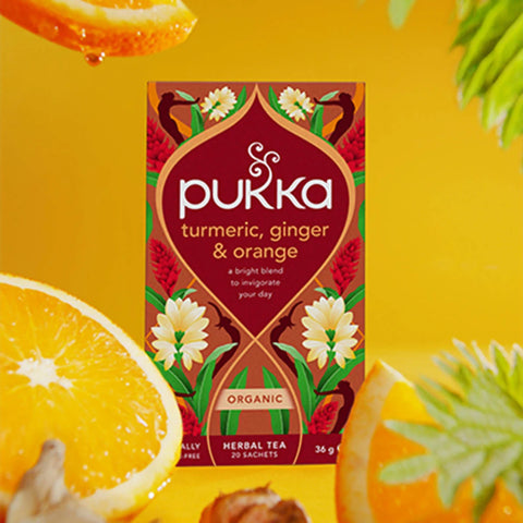 Pukka Herbs Turmeric, Ginger & Orange Tea on a yellow background with sliced orange and ginger pieces.