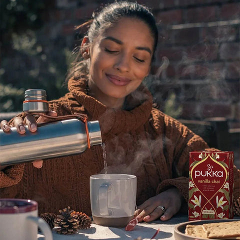 Woman in a brown sweater holding a thermos with a steaming mug, next to a box of Pukka Vanilla Chai.