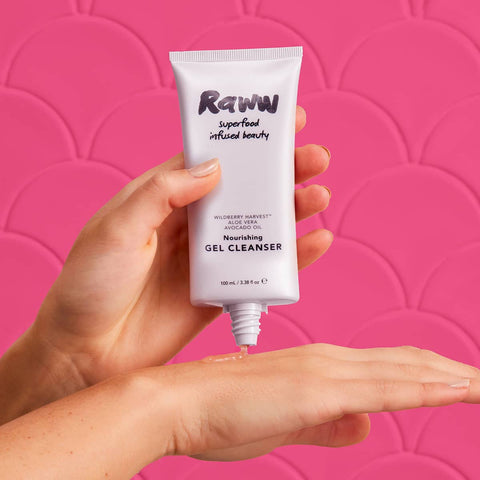Hand holding a tube of Raww Nourishing Gel Cleanser 100ml against a pink background.