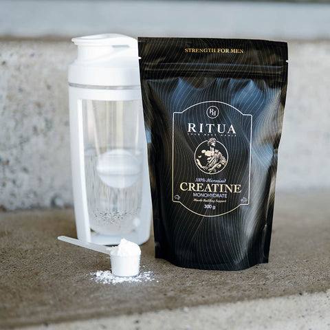 Scoop of white powder next to black packaging of Ritua Creatine - Strength For Him 300g and white shaker bottle.