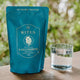 Ritua L-Glutamine packet next to a glass of water on a wooden surface with greenery in the background.