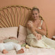 Woman sitting on a bed with a child, both wearing light-colored clothing, in a room with a wicker headboard.