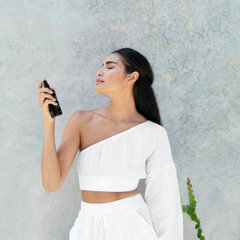 Model spraying rose mist on her face. Pictured against a marble wall with a vine climbing up. 