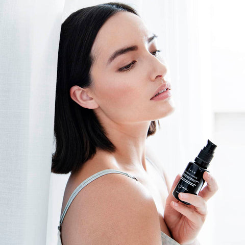 Woman leaning against a sheer white curtain holding a bottle of calming serum. 