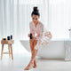 White bathroom with sheer white curtains and stool with Sodashi products. Model applying the body oil to her legs while seated on bath edge.