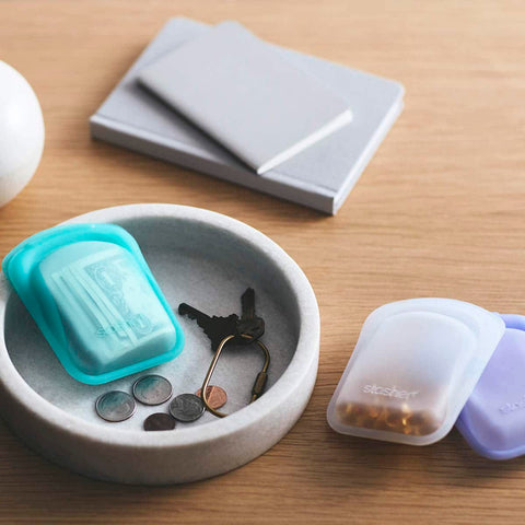 Small stone dish with keys and an aqua pocket with change inside. Sitting on a wooden surface with other stasher products.
