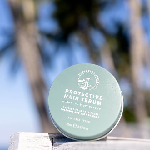 70ml tin of SunButter Skincare Protective Hair Serum with rosemary and grapeseed, set against a blurred background of palm trees. The blue tin features simple white text and is designed to nourish and protect hair from chlorine and salt damage. Suitable for all hair types.