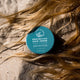 Tin of protective hair serum styled on top of a person's hair lying down in the sand.