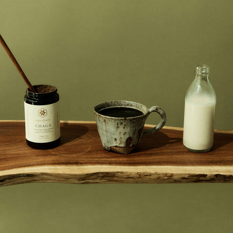 Ceramic mug with dark liquid, jar labeled 'Chaga', and bottle of milk on a wooden surface against a green background