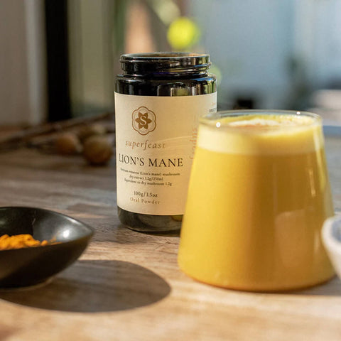 Superfeast Lion's Mane supplement jar on a wooden surface with a glass of yellow liquid.