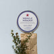 Tin of 'Miracle Manuka' cream on a stone surface with manuka twigs against a light blue background.