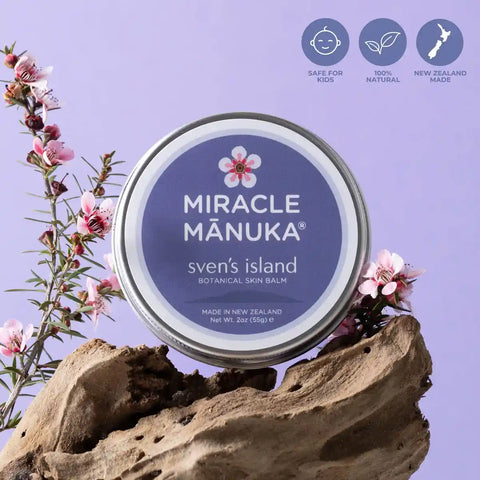Tin of Miracle Manuka balm on a piece of driftwood with manuka flowers against a purple background