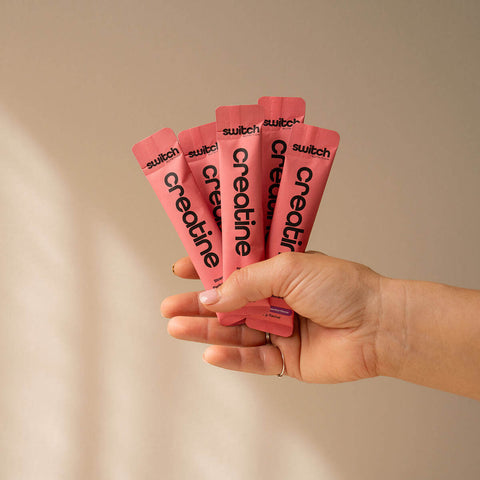 Hand holding pink packets of switch creatine against a beige background.