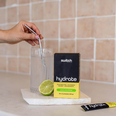 Person stirring a glass of water with a lime wedge, next to 'switch' hydrate packets on a tiled countertop.