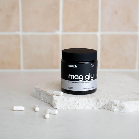 Container of mag gly supplement on a white surface with a tiled wall background.