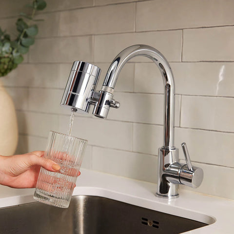 Person filling a glass with water from a kitchen faucet with a water filter attachment.