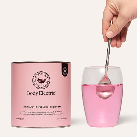 Pink protein water in a glass with a hand   and spoon beside a ‘Body Electric’ container.