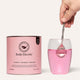 Pink protein water in a glass with a hand   and spoon beside a ‘Body Electric’ container.