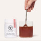 The Beauty Chef Clear Skin 150g product jar with a glass of red liquid & spoon on a white background.