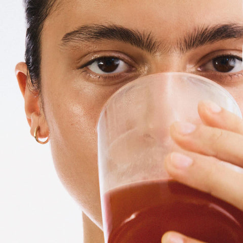 Person holding a glass of red juice close to their face on a white background.