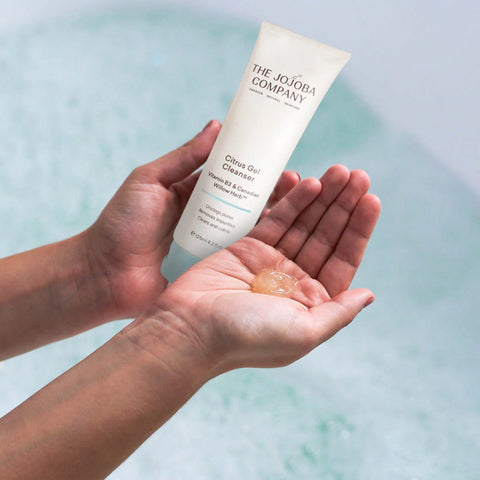 Person holding a tube of 'The Jojoba Company' Citrus Gel Cleanser with a small amount on their palm against a light blue background.
