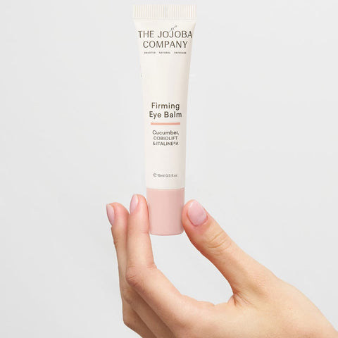 The Jojoba Company Firming eye balm tube held by a hand on a light gray background.