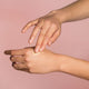 Hand applying cream to another hand against a pink background.