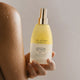 A hand holding a bottle of The Jojoba Company Jojoba Firming Body Treatment, with a person's arm visible on the left against a soft neutral background.