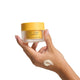 Hand holding a jar of 'The Jojoba Company' balm with a visible cream swatch on skin.