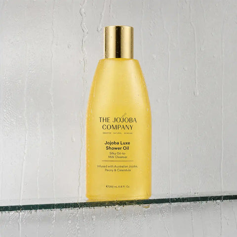 A bottle of The Jojoba Company Jojoba Luxe Shower Oil, filled with golden yellow liquid and topped with a metallic gold cap, resting on a glass shelf against a textured, rain-slicked gray background.