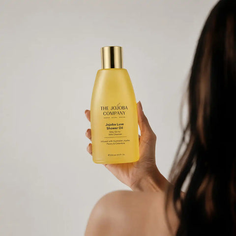 A hand holds a bottle of The Jojoba Company Jojoba Luxe Shower Oil against a neutral light gray background, with the back of a woman's head and shoulder visible on the right.
