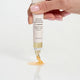 A hand with pale pink manicured nails holds a white tube of lip treatment above a glossy white surface, with the product squeezed out as a small amber-colored droplet.