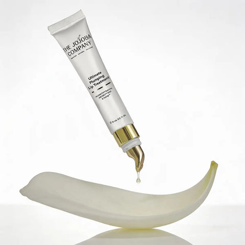 A white tube of The Jojoba Company Ultimate Plumping Lip Treatment with a gold cap, shown dispensing a drop of cream onto a curved white dish against a clean white background.