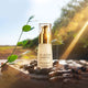 The Jojoba Company Ultimate Serum sitting on a wooden table with jojoba seeds scattered around and trees in the background with a ray of sunshine highlighting the serum.