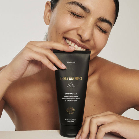 Woman holding a 'Three Warriors' 'Gradual Tan' product against a neutral background.