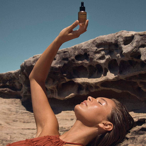 Woman lying on a rock with a bottle of tan drops held above her. Blue sky bronze tones.