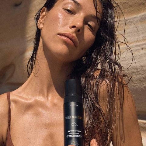 Tan model in a brown bathing suit with a bottle of self-tan mousse at a beach with wet hair.