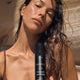 Tan model in a brown bathing suit with a bottle of self-tan mousse at a beach with wet hair.