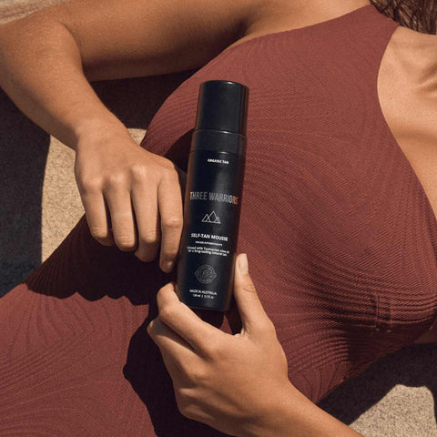 Close up of model laying on the beach in a red-brown swimsuit holding three warriors self tan mousse.