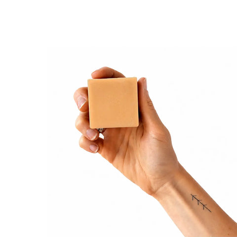 A hand holding a square, light tan bar of soap against a solid white background.