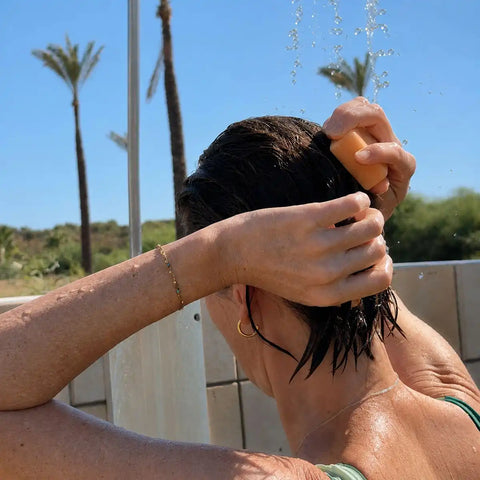 A woman outdoors under a shower stream washes her dark hair with an orange soap bar. Palm trees and a blue sky are visible in the background.