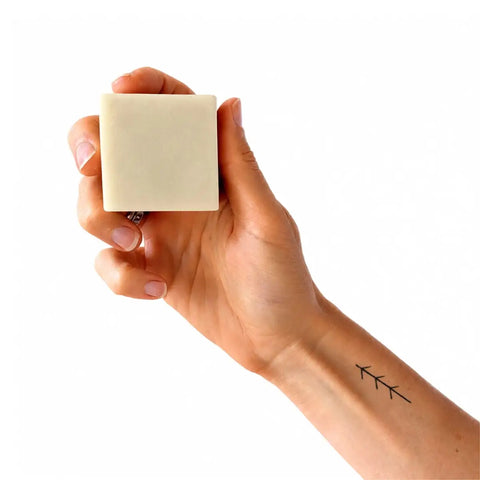 A hand holding a small, square, off-white bar of soap against a solid white background.