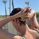 A woman outdoors under a shower, washing her hair with a pale yellow shampoo bar. She is shown from behind, with water spraying down, palm trees, and a blue sky in the background.