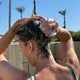 A woman outdoors with wet dark hair lathered in shampoo, shown from the back against a sunny blue sky with palm trees and in the background.