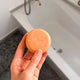 A hand holding a round, orange-colored soap bar in front of a white bathtub.