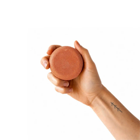 Hand holding a round, terracotta-coloured soap bar against a white background.