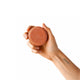 Hand holding a round, terracotta-coloured soap bar against a white background.